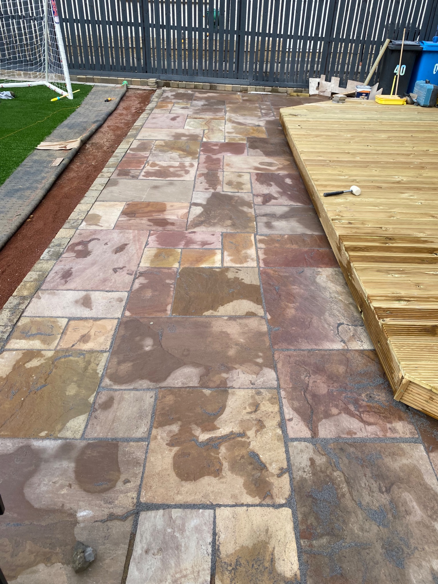 Świeżo ułożona, mokra, wielobarwna kamienna ścieżka ogrodowa z jasnymi spoinami, granicząca z trawnikiem ze sztucznej trawy i częściowo ukończonym drewnianym tarasem, widoczne narzędzia pracy...