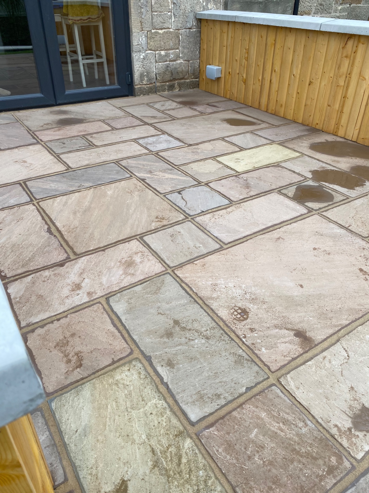 Ułożona, wielobarwna kamienna posadzka tarasu z widocznymi spoinami, fragment drewnianej balustrady i ślady wilgoci na powierzchni