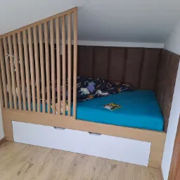 Łóżko dziecięce w zabudowie antresoli z drewnianą barierką z pionowych listewek, tapicerowanym wezgłowiem i wysuwanymi szufladami na pościel, w pokoju na poddaszu ze skośnym sufitem.