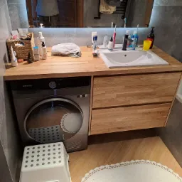 Łazienka z drewnianym blatem, pod którym umieszczono pralko-suszarkę Samsung. Nad blatem umywalka i lustro z oświetleniem LED. Na blacie kosmetyki i akcesoria łazienkowe.