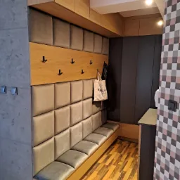 Wnętrze przedpokoju z zabudową na wymiar: tapicerowany panel ścienny z siedziskiem, drewniane wieszaki i szafki pod sufitem, ciemnoszara zabudowa w głębi, podłoga z paneli imitujących drewno.