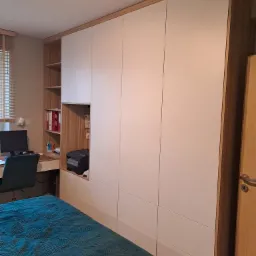 Duża, biała szafa wnękowa z drewnianymi akcentami, wkomponowana w przestrzeń pokoju z biurkiem i łóżkiem; widoczny fragment blatu biurka z komputerem i regał z segregatorami.