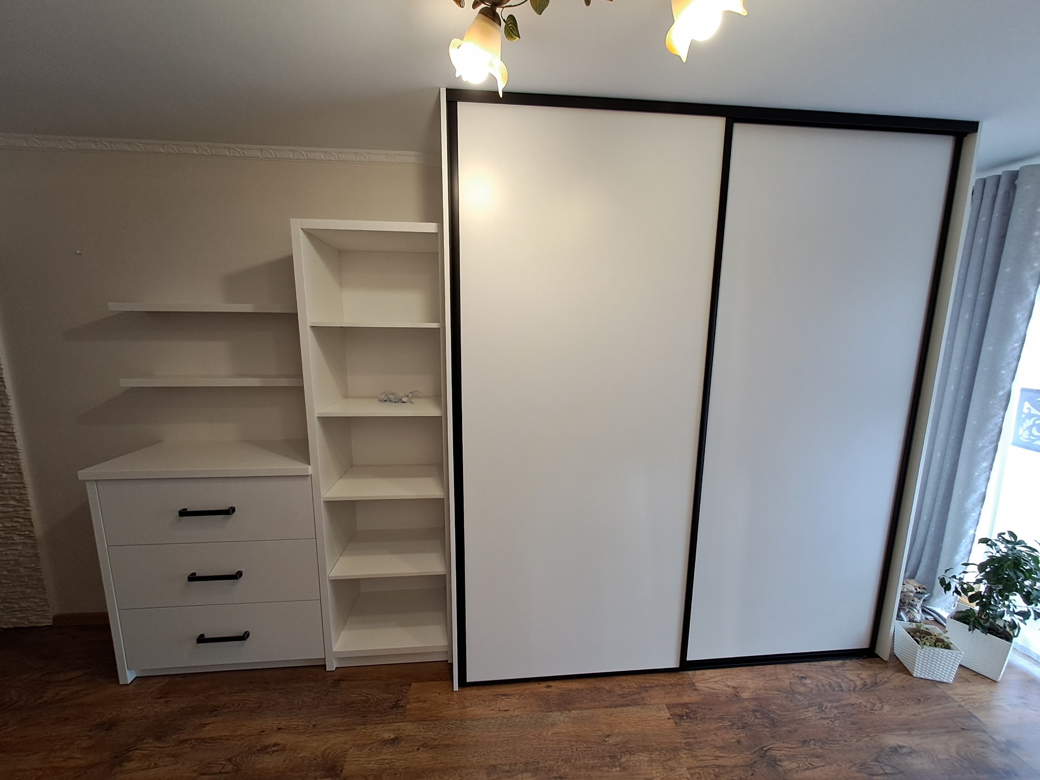 Biała szafa przesuwna z czarnymi obramowaniami, po lewej stronie biała komoda z trzema szufladami i regał z półkami, na podłodze panele, obok szafy doniczki z kwiatami.