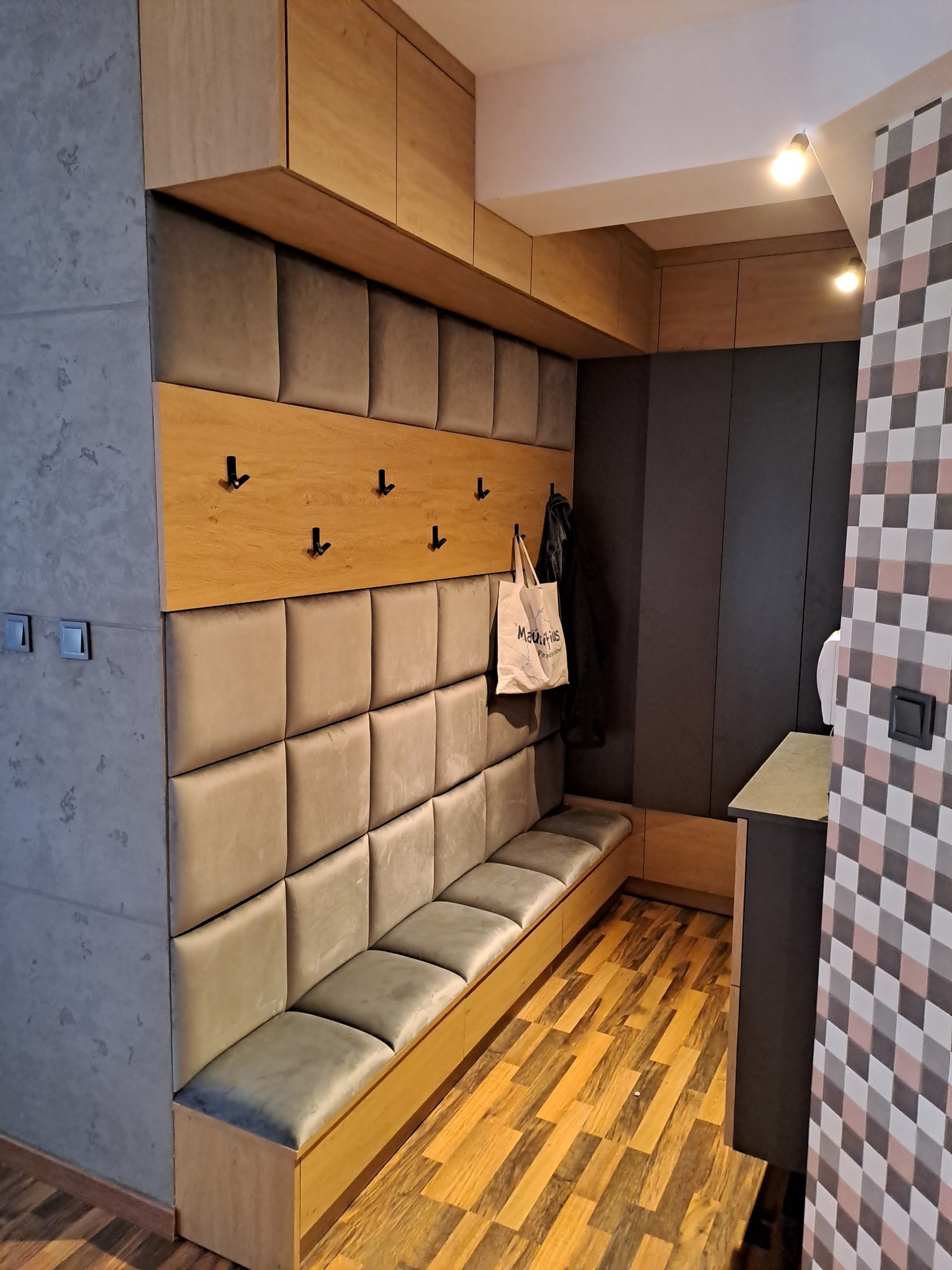 Wnętrze przedpokoju z zabudową na wymiar: tapicerowany panel ścienny z siedziskiem, drewniane wieszaki i szafki pod sufitem, ciemnoszara zabudowa w głębi, podłoga z paneli imitujących drewno.