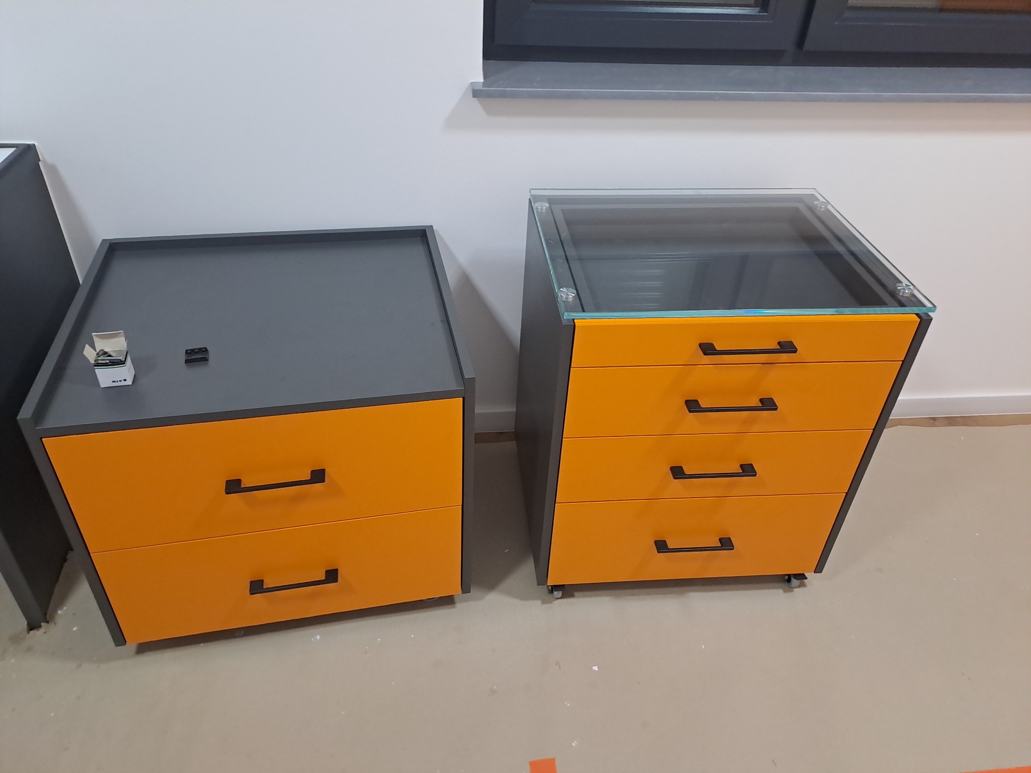 Dwie szafki z szufladami: jedna z blatem szarym, druga z blatem szklanym, obie w kolorze pomarańczowym z czarnymi uchwytami, na podłodze w surowym stanie.