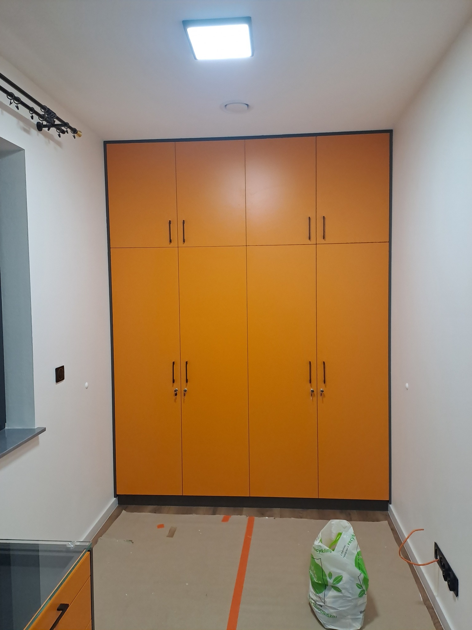 Pomarańczowa szafa wnękowa z czarnymi obramowaniami i uchwytami, widoczna w trakcie montażu, podłoga zabezpieczona kartonem, worek na śmieci obok gniazdka elektrycznego.