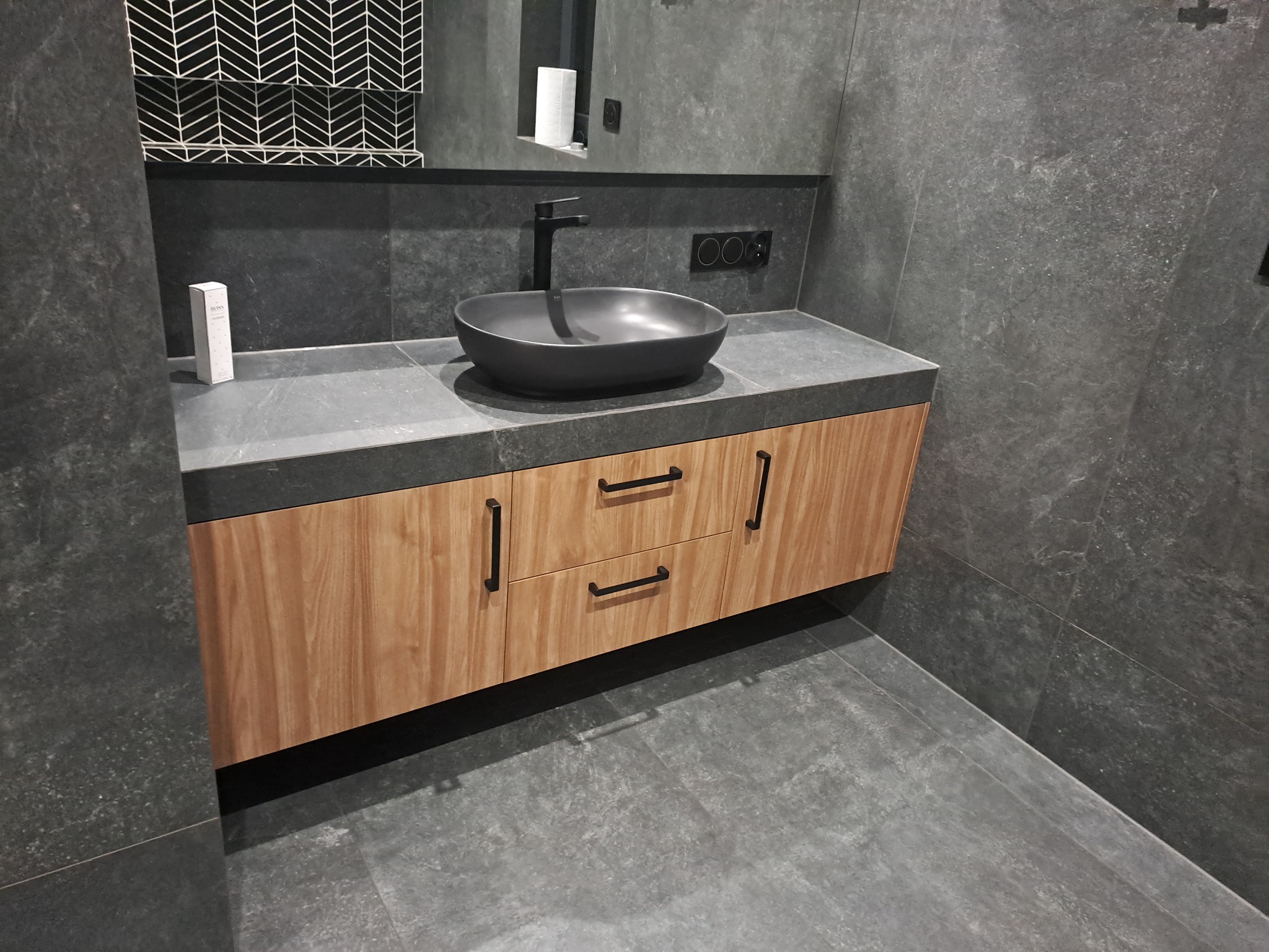 Łazienka z drewnianą szafką pod umywalką nablatową w kolorze grafitowym, czarna armatura i szara płytka imitująca kamień na ścianach i podłodze.