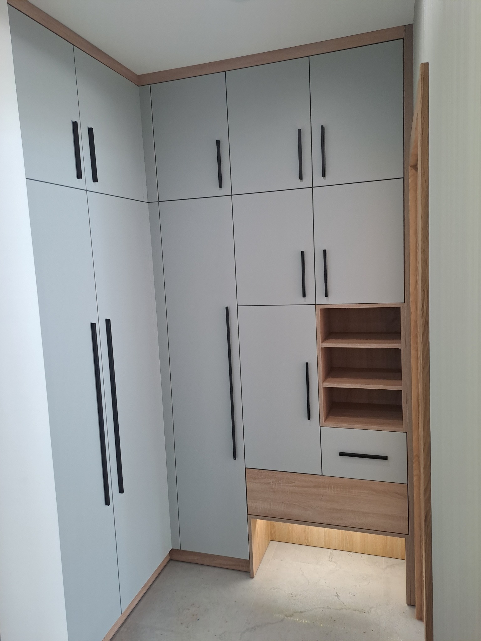 Jasnoszara szafa wnękowa z drewnianymi akcentami i czarnymi uchwytami, oświetlenie LED u dołu, widok z dołu.