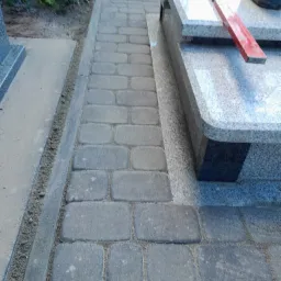 Świeżo ułożona ścieżka z szarej kostki brukowej prowadząca do pomnika na cmentarzu, z poziomnicą leżącą na płycie nagrobnej.