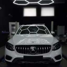 Biały samochód marki Mercedes z nowym grillem, stojący w ciemnym pomieszczeniu z podświetlanym heksagonalnym oświetleniem sufitowym, widziany z przodu.