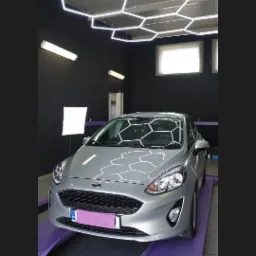 Srebrny Ford Fiesta na fioletowym podnośniku w czarnym pomieszczeniu z heksagonalnym oświetleniem sufitowym.