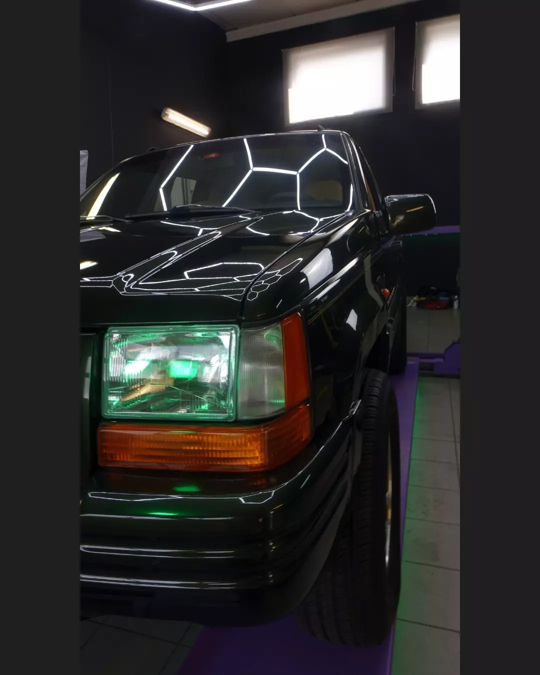 Czarny samochód Jeep Cherokee po detailingu, z wyraźnie odbijającym się światłem na lakierze i zielonym podświetleniem reflektorów, ustawiony w sterylnym, ciemnym pomieszczeniu z fioletową podłogą.