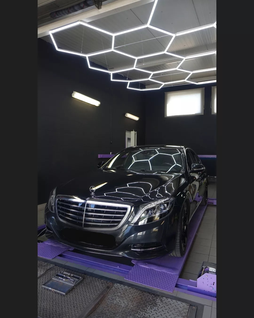Czarny Mercedes klasy S na podnośniku w warsztacie z heksagonalnym oświetleniem sufitowym; skrzynka z narzędziami widoczna obok podnośnika.