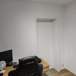 Pokój z biurkiem, monitorem, wentylatorem i krzesłem gamingowym Pro-Gamer przed otwartymi drzwiami, parkiet na podłodze.