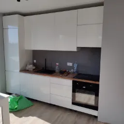 Nowoczesna, biała kuchnia z drewnianym blatem i czarnym zlewem, widoczne narzędzia i butelka wody, sufit z czarnym oświetleniem punktowym, podłoga panelowa, w rogu zielona torba.