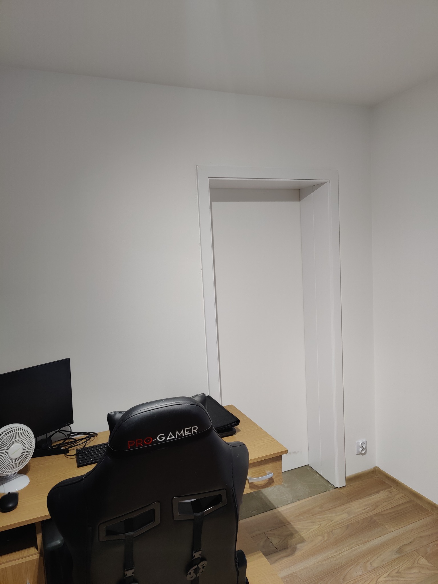 Pokój z biurkiem, monitorem, wentylatorem i krzesłem gamingowym Pro-Gamer przed otwartymi drzwiami, parkiet na podłodze.