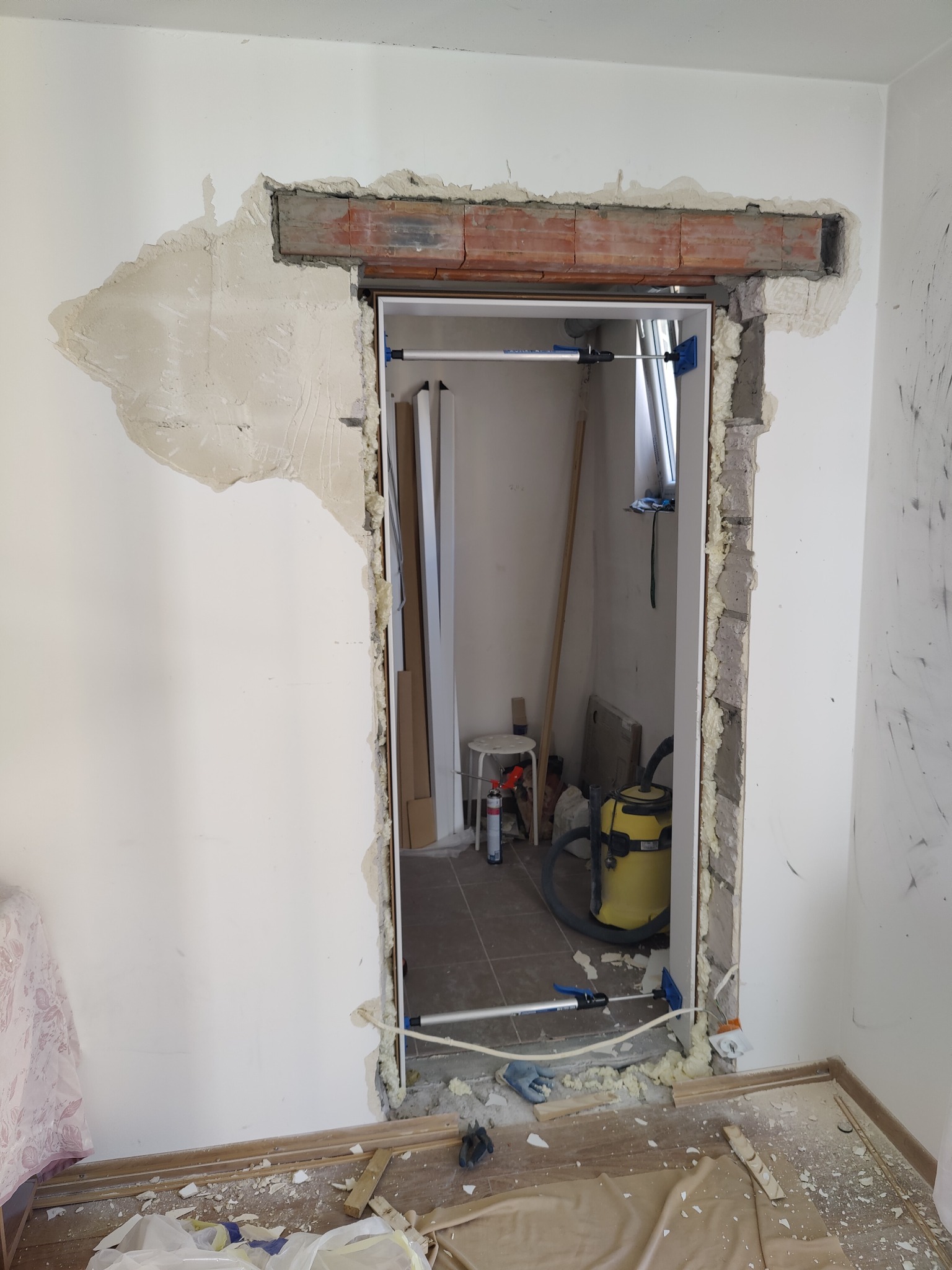 Otwór drzwiowy w trakcie poszerzania, widoczne cegły nadproża, pianka montażowa, uszkodzony tynk i narzędzia w tle.