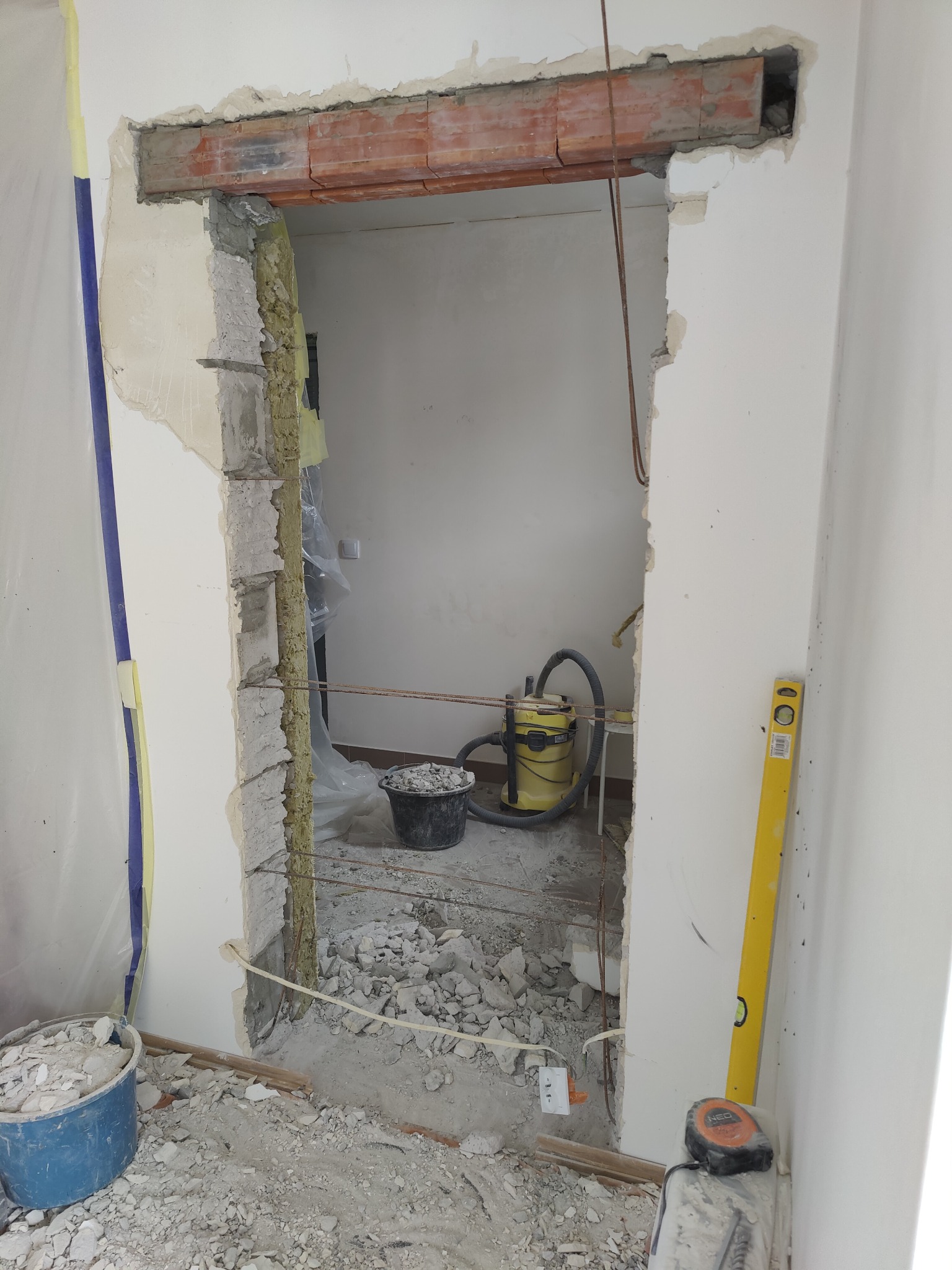 Otwór w ścianie po usunięciu futryny, widoczne cegły nadproża, gruz, wiadro i odkurzacz przemysłowy w tle.