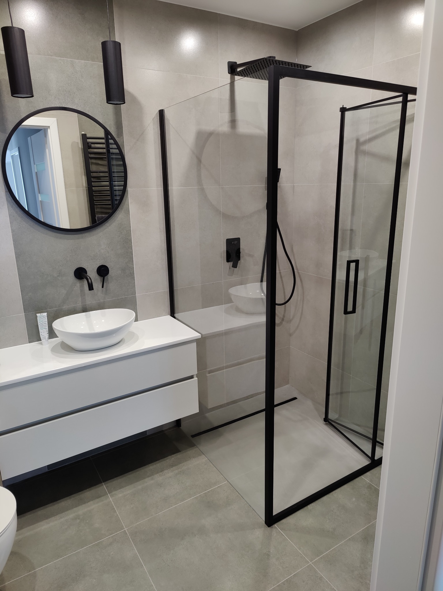 Nowoczesna łazienka z kabiną prysznicową typu walk-in z czarnymi ramami, umywalką nablatową i okrągłym lustrem z czarną ramą, oświetlona wiszącymi lampami tubami.