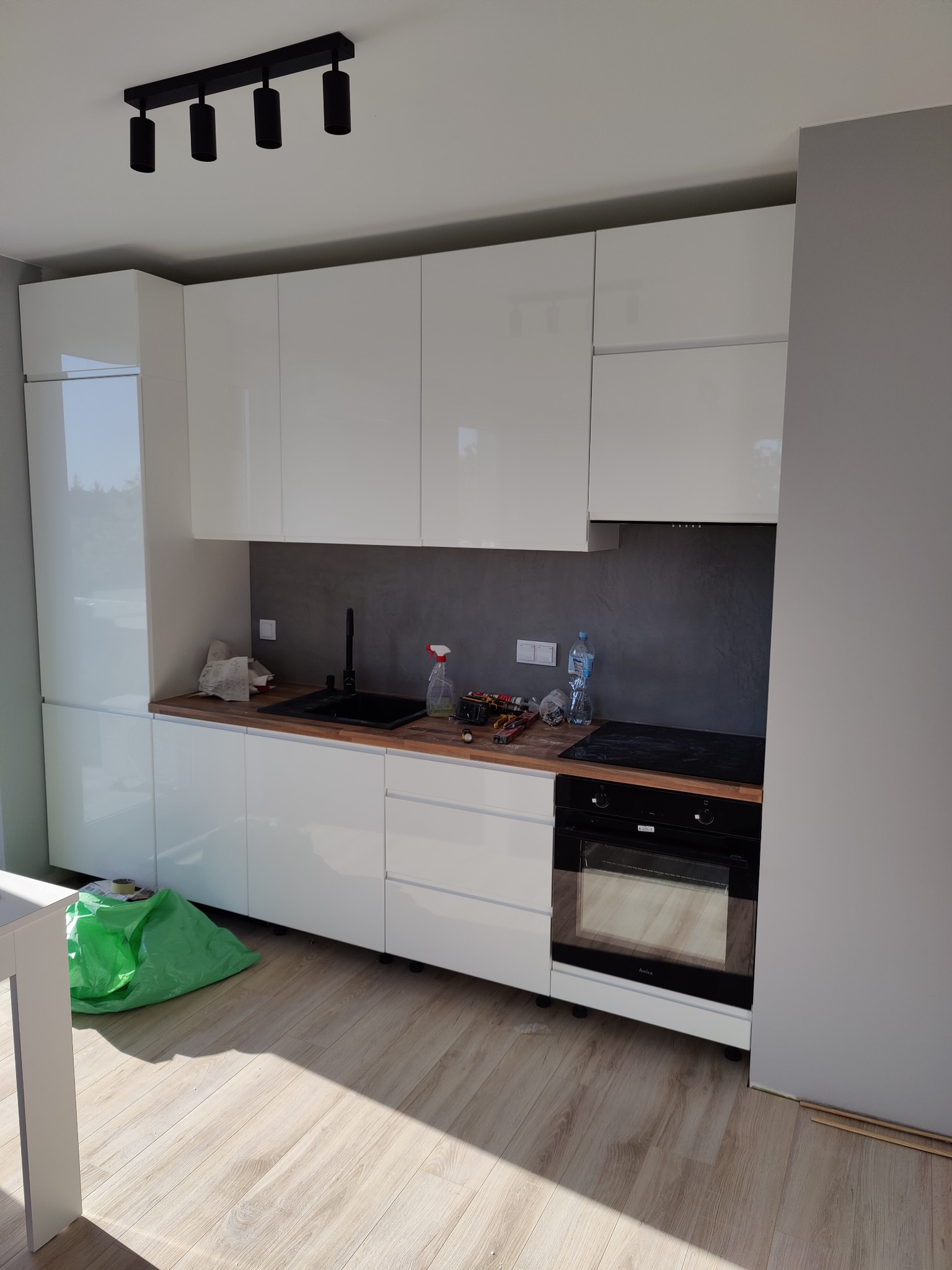 Nowoczesna, biała kuchnia z drewnianym blatem i czarnym zlewem, widoczne narzędzia i butelka wody, sufit z czarnym oświetleniem punktowym, podłoga panelowa, w rogu zielona torba.