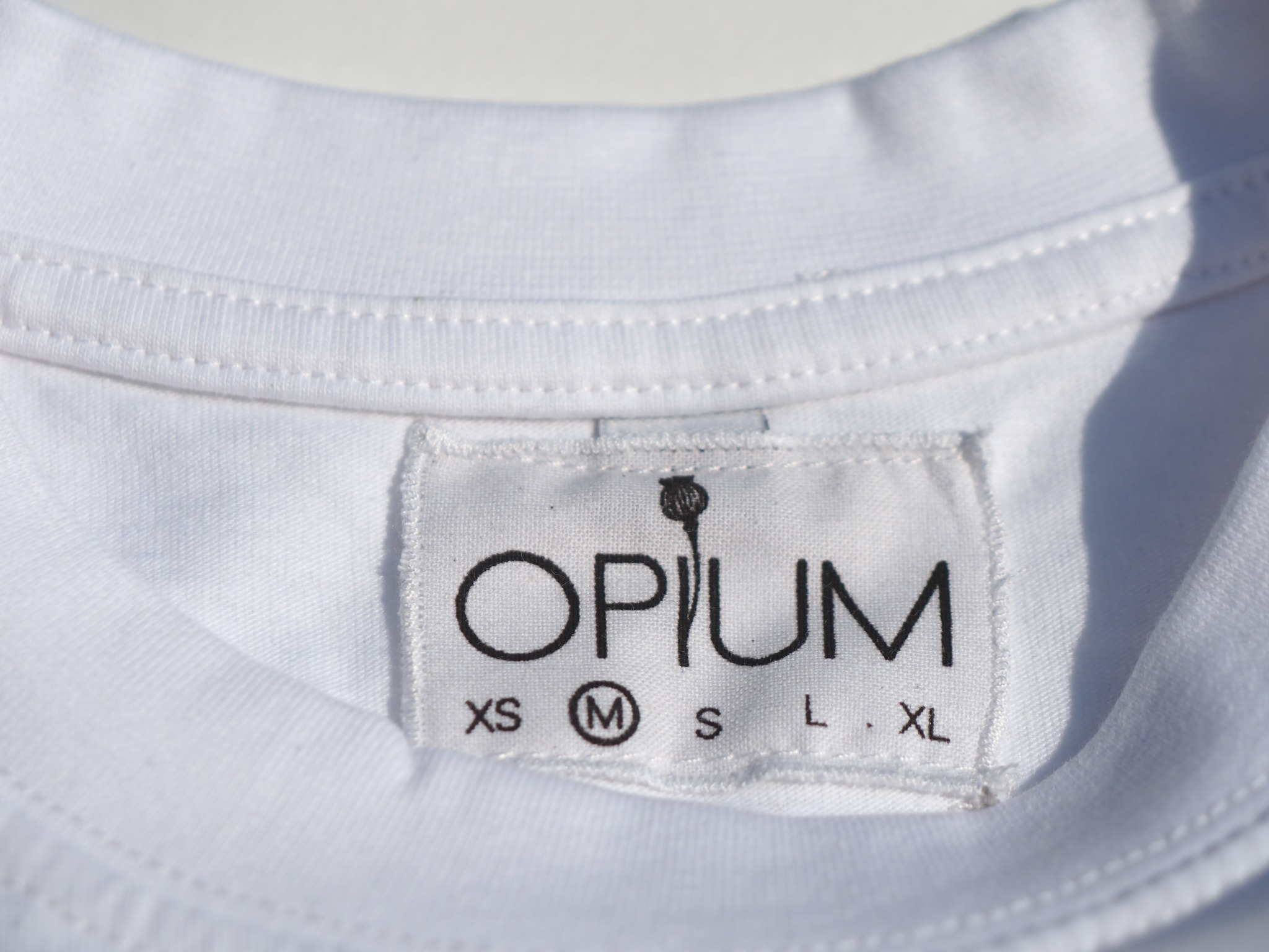 Biała koszulka z metką 'Opium' i zaznaczonym rozmiarem 'M', widoczne szwy przy dekolcie.