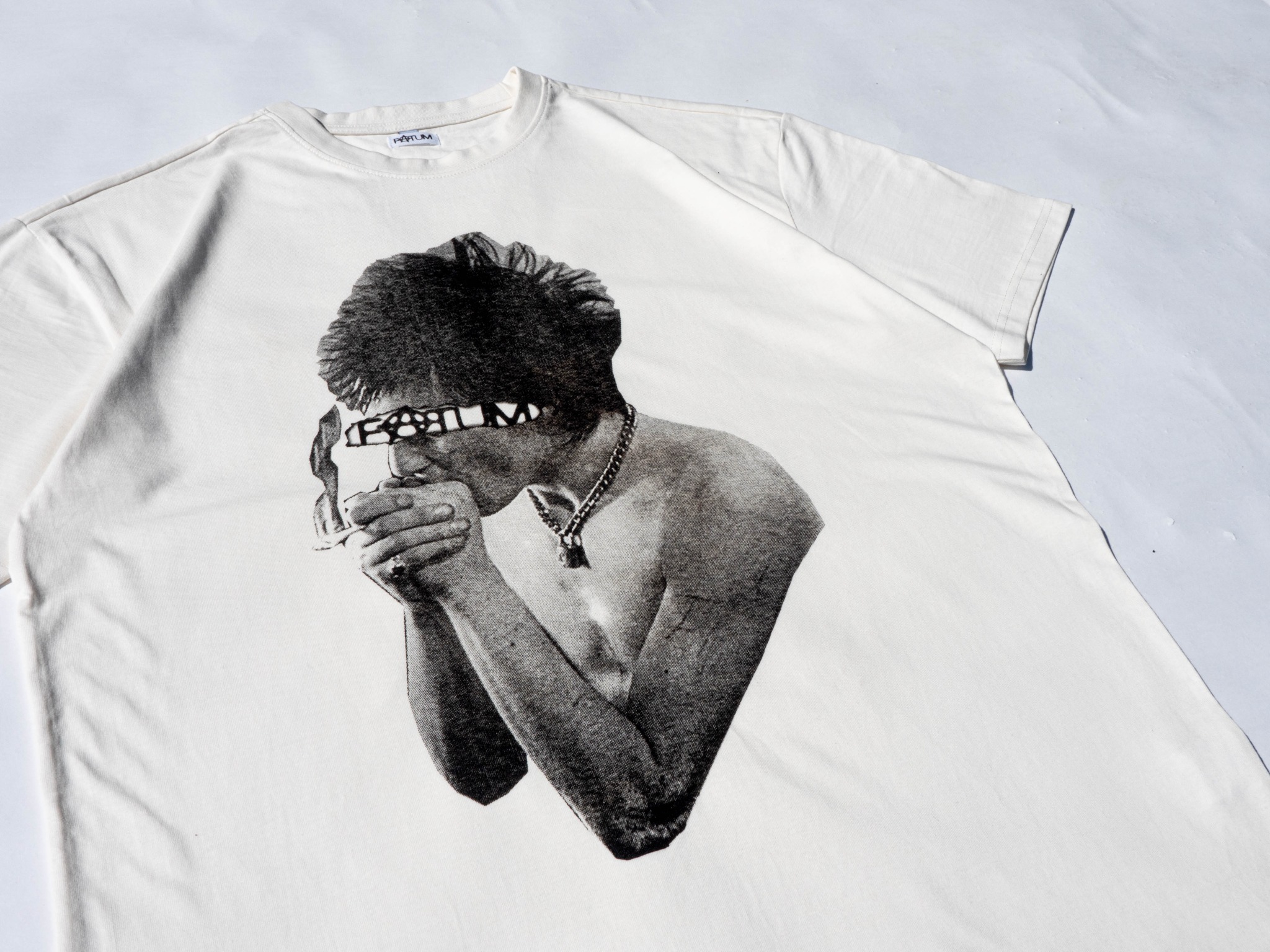 Biała koszulka z czarno-białym nadrukiem przedstawiającym mężczyznę z papierosem i łańcuchem na szyi, leżąca na białym tle, widoczne logo producenta na kołnierzyku.