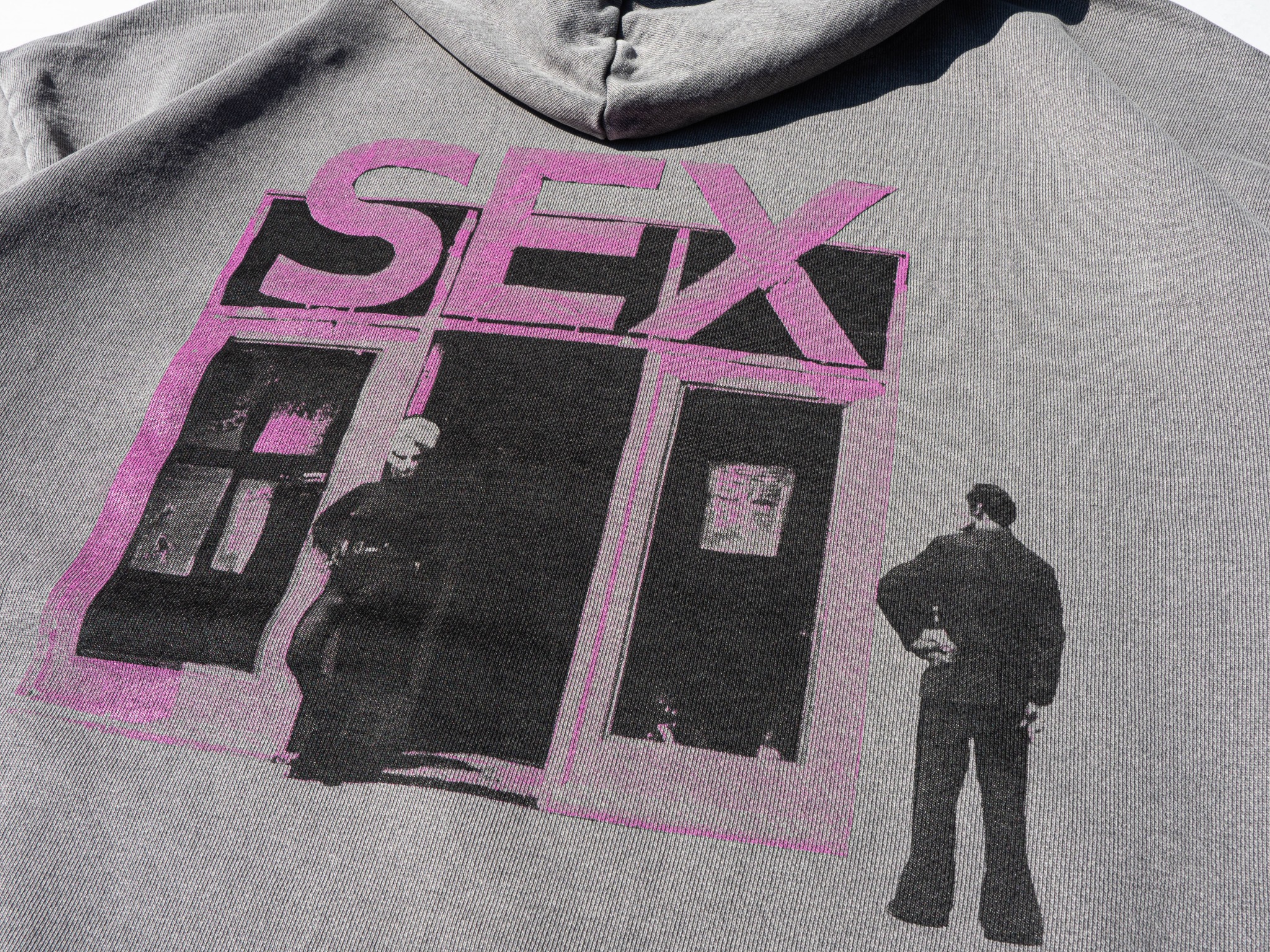 Szara bluza z kapturem, na której widnieje nadruk przedstawiający fasadę budynku z napisem 'SEX' w kolorze różowym, a także sylwetki dwóch postaci w czerni.