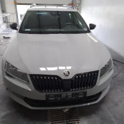 Szary samochód Skoda Superb kombi, po lakierowaniu, z widocznym grillem, reflektorami i logo, stojący w warsztacie z narzędziami do polerowania w tle.