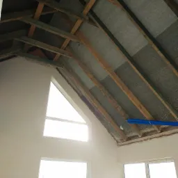 Poddasze z drewnianą konstrukcją dachu, ocieplone wełną mineralną, widoczne niebieskie rury instalacyjne, ściany w trakcie prac wykończeniowych, okna z widokiem na zewnątrz.