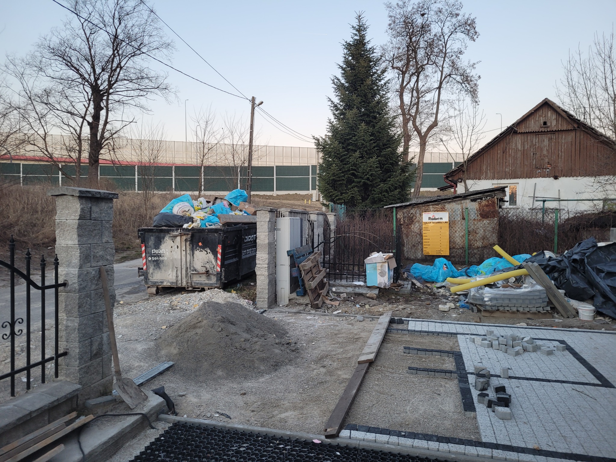 Fragment posesji w trakcie prac brukarskich: ułożona część kostki, hałda piasku, kontener na odpady budowlane i narzędzia.