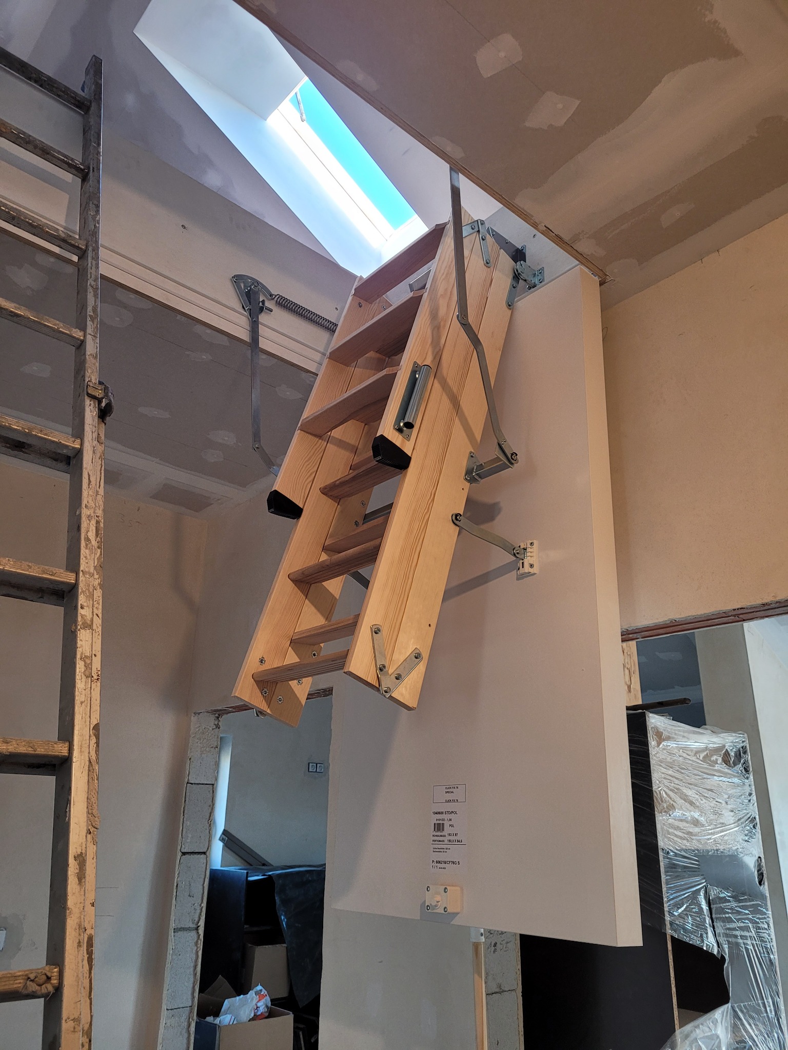 Drewniane schody strychowe składane, zamontowane w suficie, widoczne mechanizmy składania, obok stara drabina, sufit i ściany w trakcie prac wykończeniowych, w tle okno dachowe.