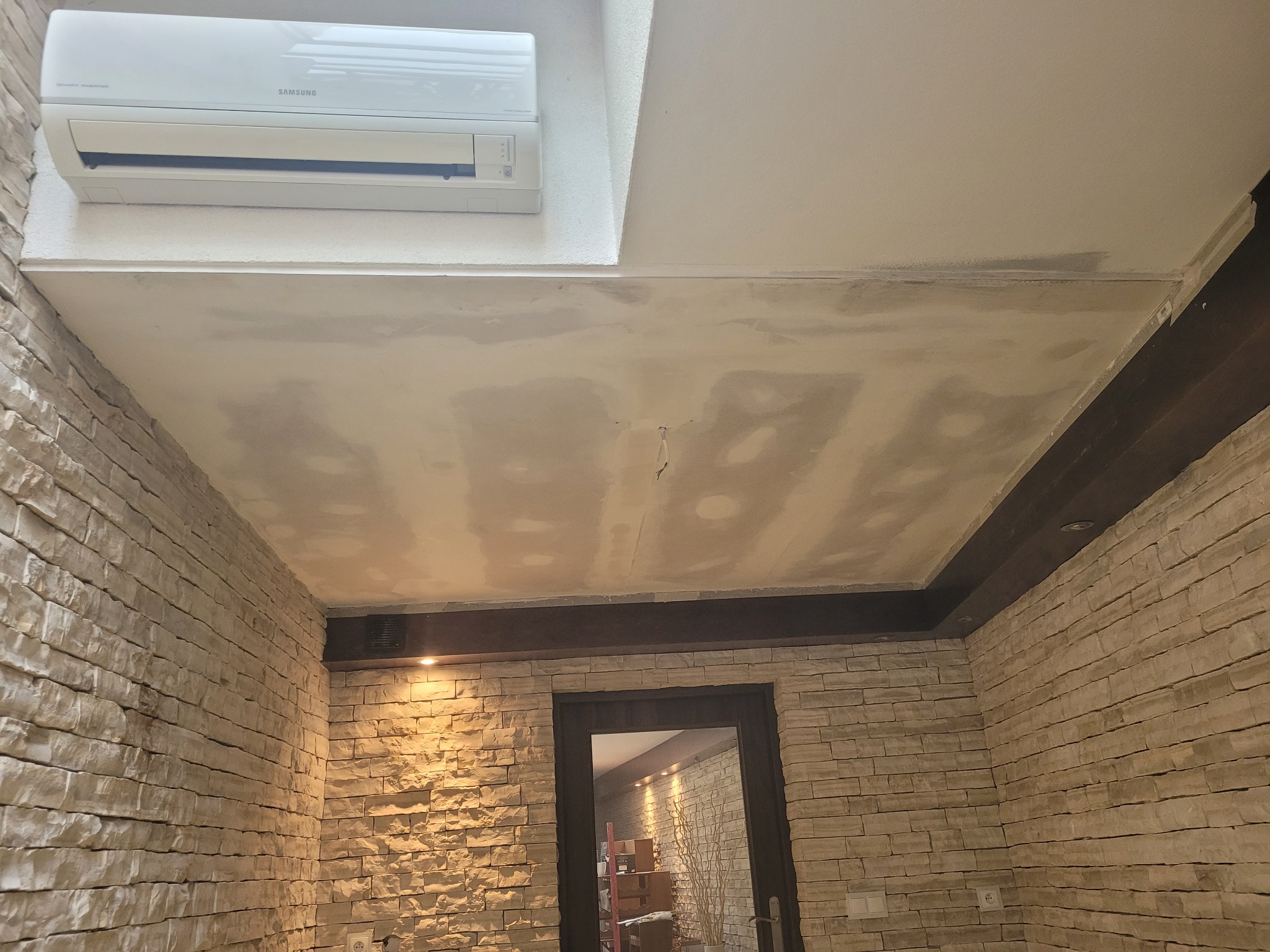 Wnętrze pokoju z kamienną okładziną ścian, sufit z widocznymi śladami szpachlowania, klimatyzator na suficie i ciemna listwa dekoracyjna pod sufitem.