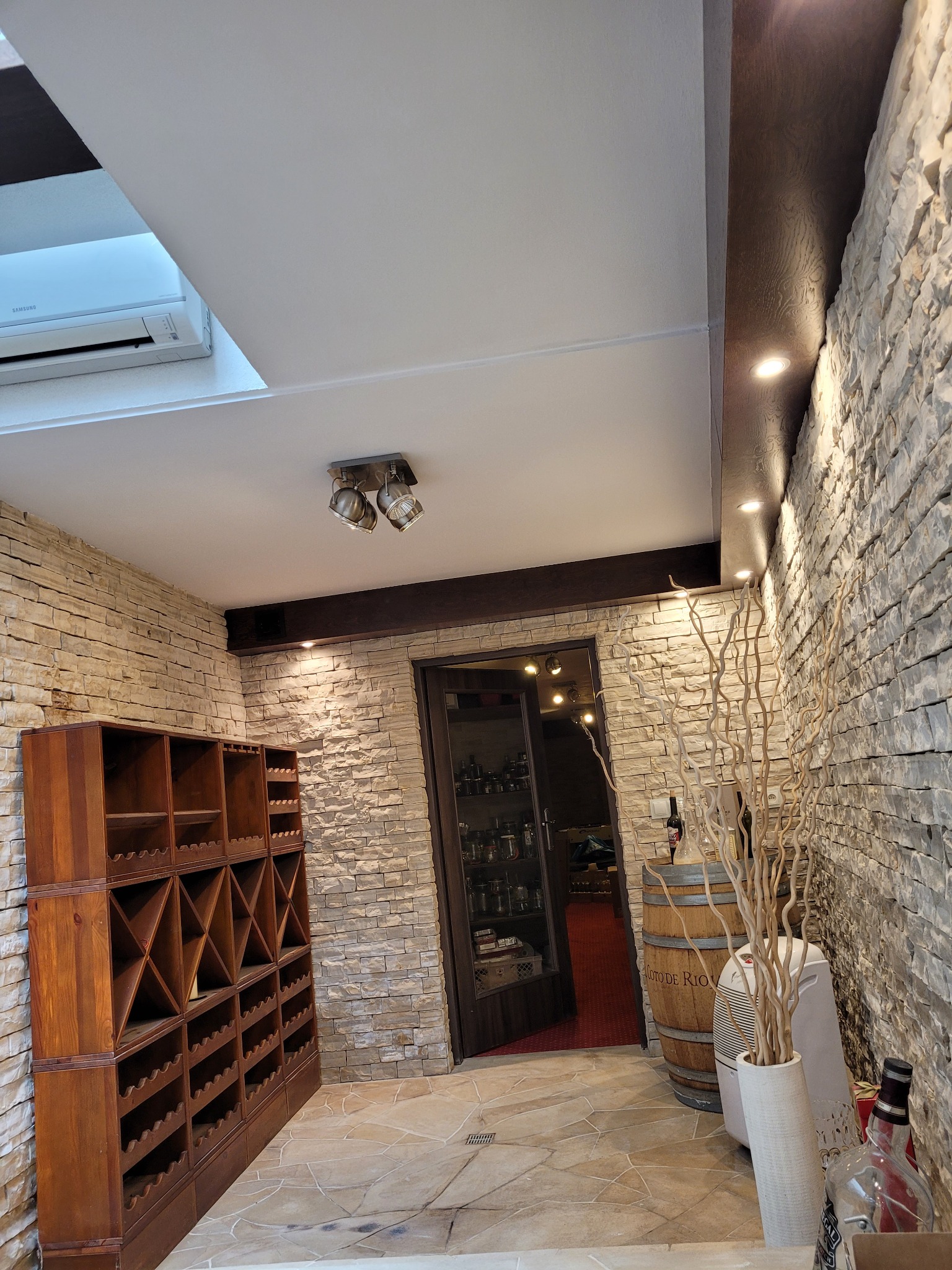 Wnętrze winiarni z kamienną ścianą, drewnianym regałem na wino, beczką i klimatyzatorem podsufitowym.