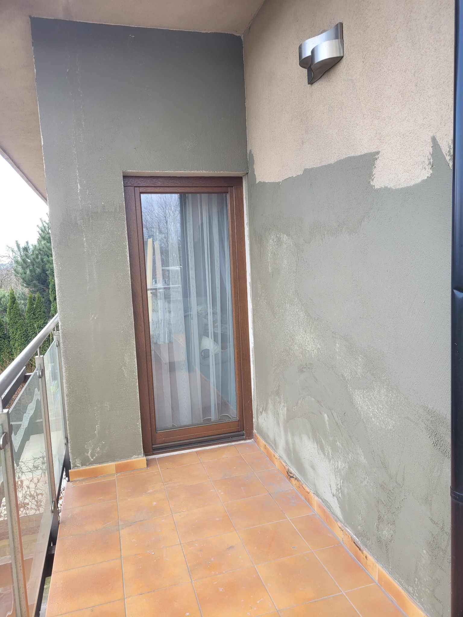 Balkon w trakcie remontu, ściana częściowo pokryta szarą warstwą, widoczne ślady po wcześniejszych naprawach, brązowe płytki na podłodze, okno balkonowe z drewnianą ramą.