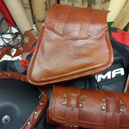 Usługi kaletnicze i szewskie - Brązowe, skórzane akcesoria motocyklowe: torba, sakwa i element siedzenia z nitami, leżące na czarno-czerwonej kurtce motocyklowej.