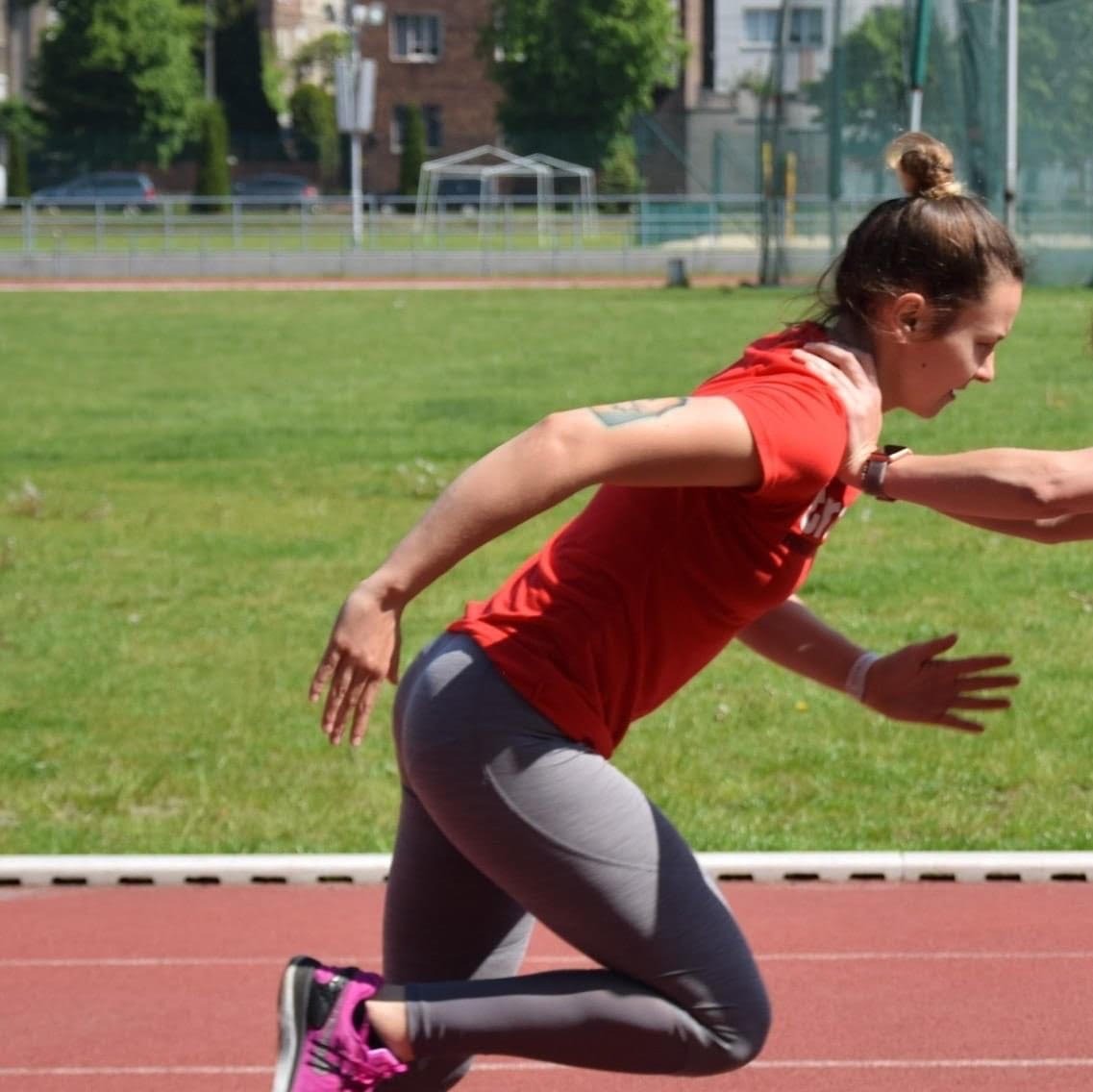 Kobieta w czerwonej koszulce i szarych legginsach biegnie po czerwonej bieżni, druga osoba pomaga jej w starcie, widoczne zielone boisko w tle.