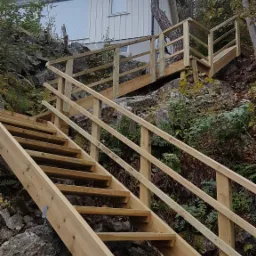 Nowe drewniane schody zewnętrzne prowadzące do domu na zboczu wzgórza, otoczone skałami i zielenią.