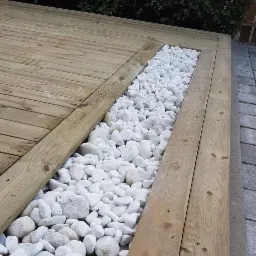 Drewniany taras z jasnego drewna z wbudowanym korytem wypełnionym białymi, otoczakami.