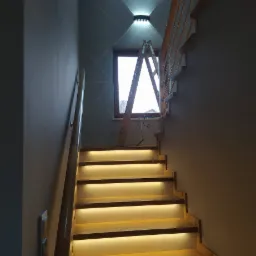 Drewniane schody z podświetleniem LED na każdym stopniu, drabina oparta o ścianę z zamontowanym kinkietem LED, instalacja w trakcie realizacji.