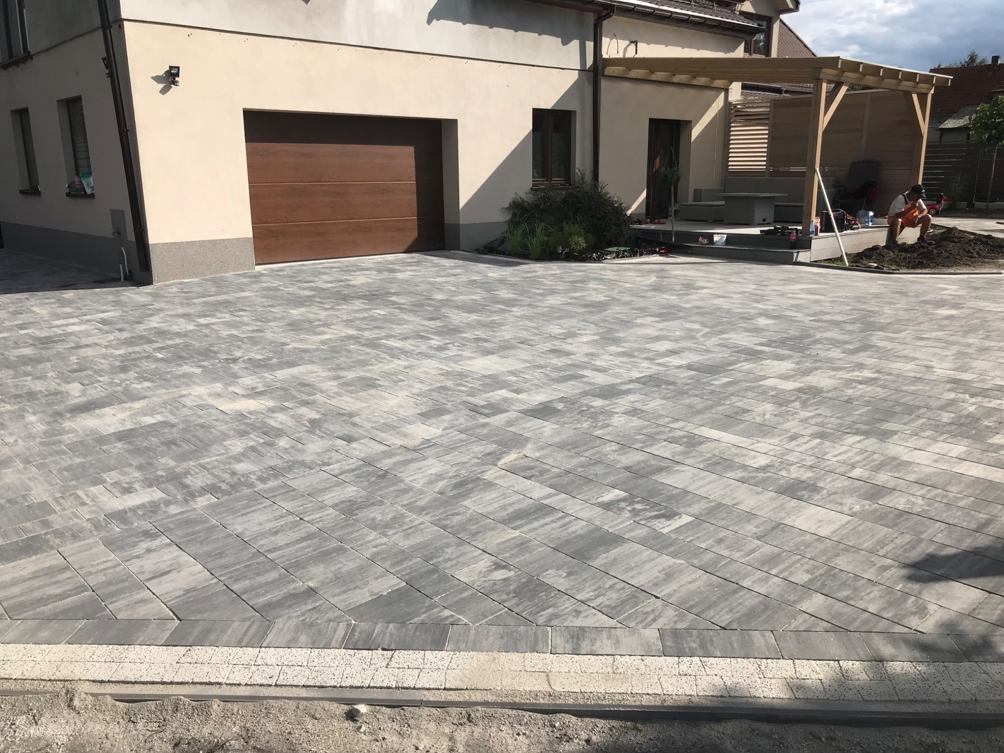 Ułożona szara kostka brukowa na podjeździe przed domem z brązową bramą garażową, widoczna drewniana pergola i pracownik w pomarańczowych spodenkach.