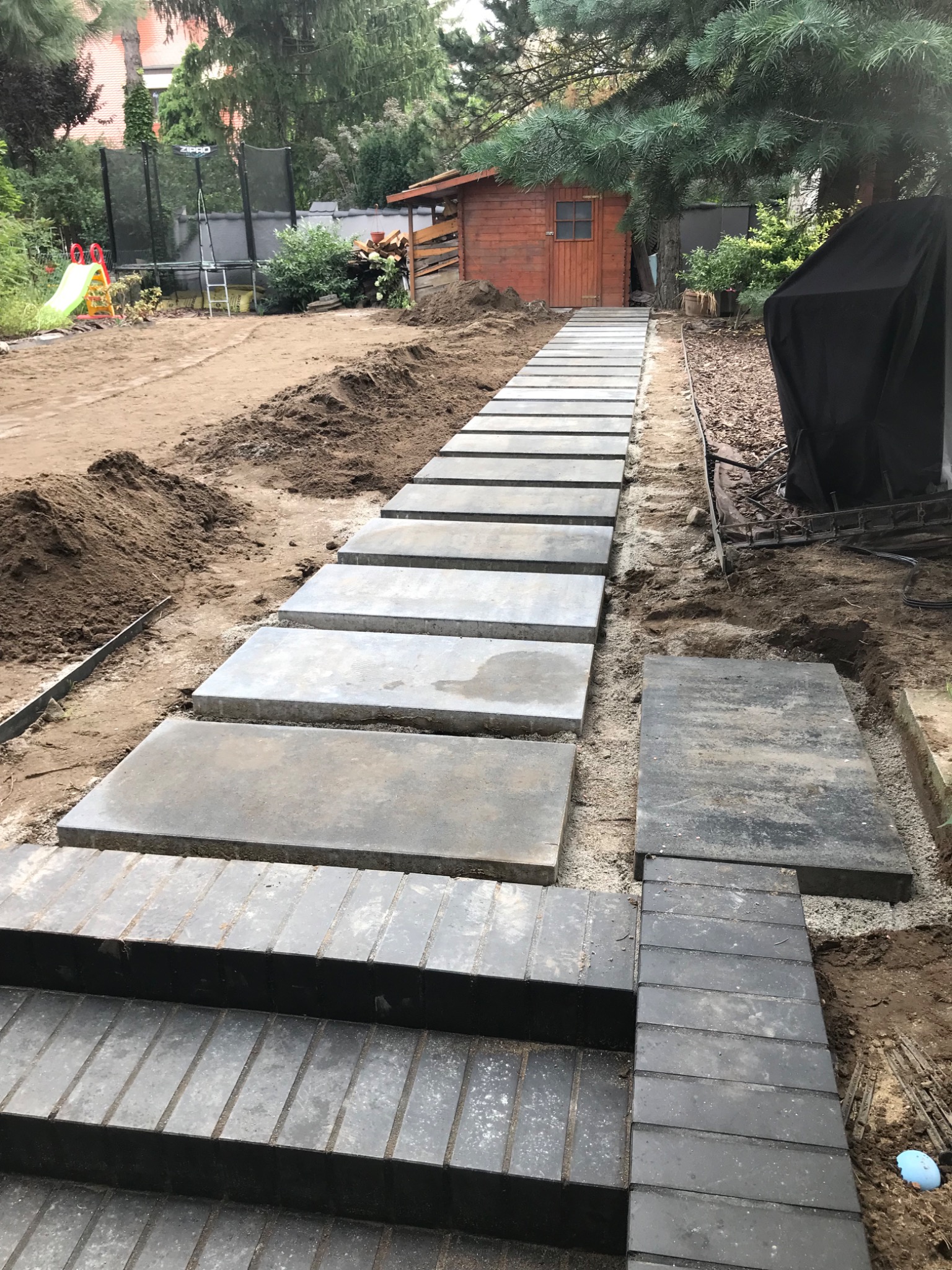 Ścieżka z betonowych płyt i kostki brukowej w ogrodzie, widoczne etapy prac ziemnych i przygotowania podłoża, w tle domek narzędziowy i zieleń