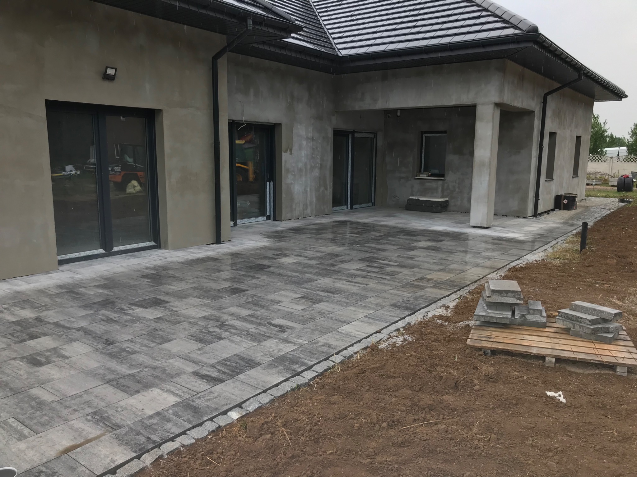 Ułożona szara kostka brukowa na podjeździe domu w stanie surowym, z paletą kostek obok, widok w pochmurny dzień.