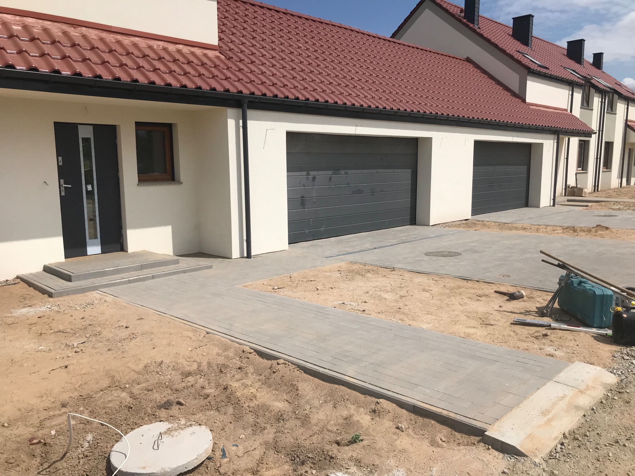 Świeżo ułożona, szara kostka brukowa prowadząca do wejścia i garażu szeregowego domu w Miękini, widoczne narzędzia brukarskie i przygotowany teren pod dalsze prace.