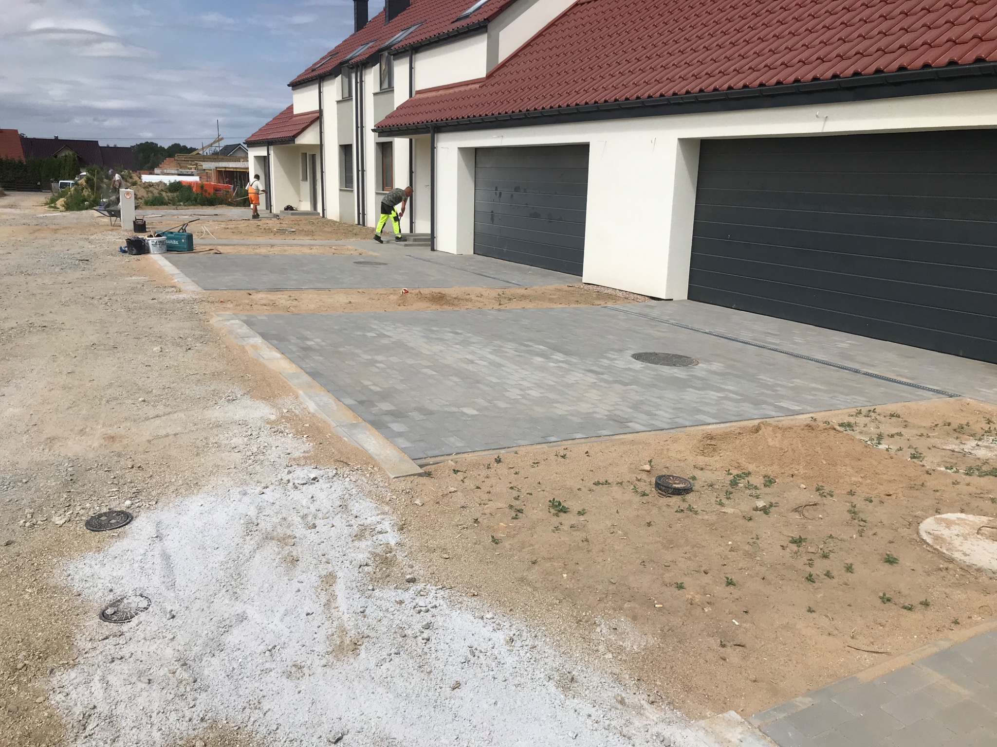 Układanie szarej kostki brukowej na podjeździe przed szeregiem nowych domów z garażami; widoczni pracownicy w odblaskowych ubraniach.