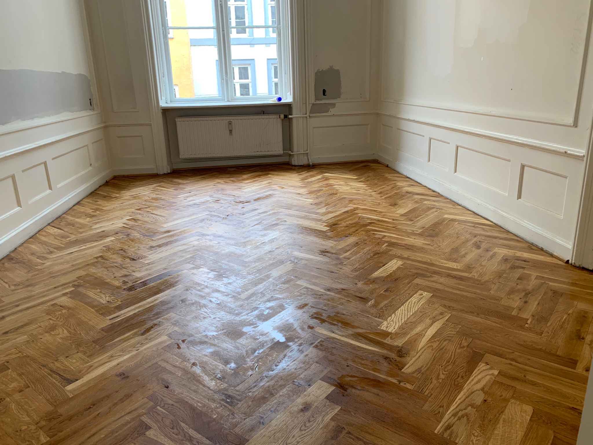 Pokój z parkietem w jodełkę po cyklinowaniu i lakierowaniu, widoczne niedomalowane fragmenty ścian i listew przypodłogowych.