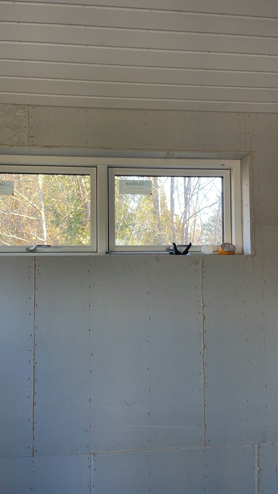 Wnętrze w trakcie prac wykończeniowych: ściana z płyt, widoczne łączenia wypełnione masą, okno z widokiem na drzewa, narzędzia na parapecie i sufit z paneli.