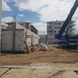 Wznoszenie ścian z białych bloczków na placu budowy wielorodzinnego budynku, widoczny żuraw budowlany i zbrojenia.