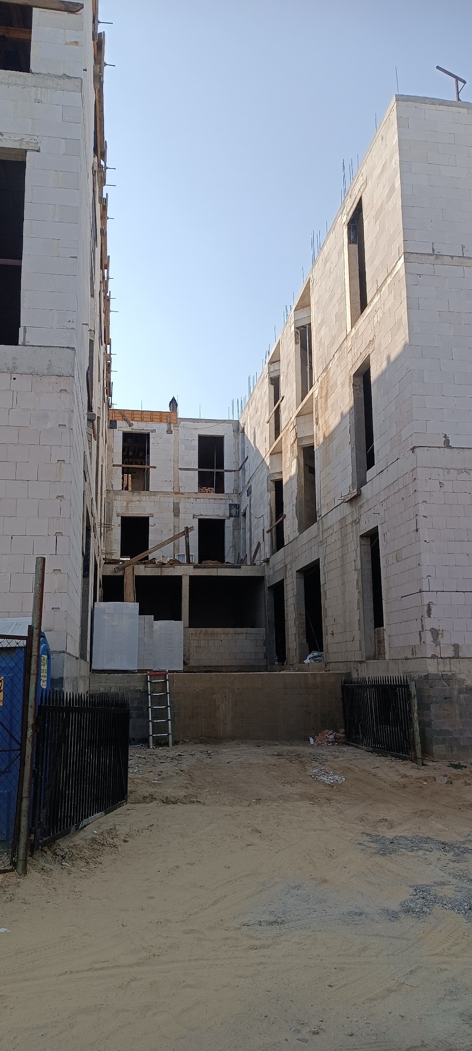 Wnętrze placu budowy z widokiem na surowe, trzypiętrowe budynki z białych bloczków, widoczne rusztowania i drabina, teren piaszczysty.