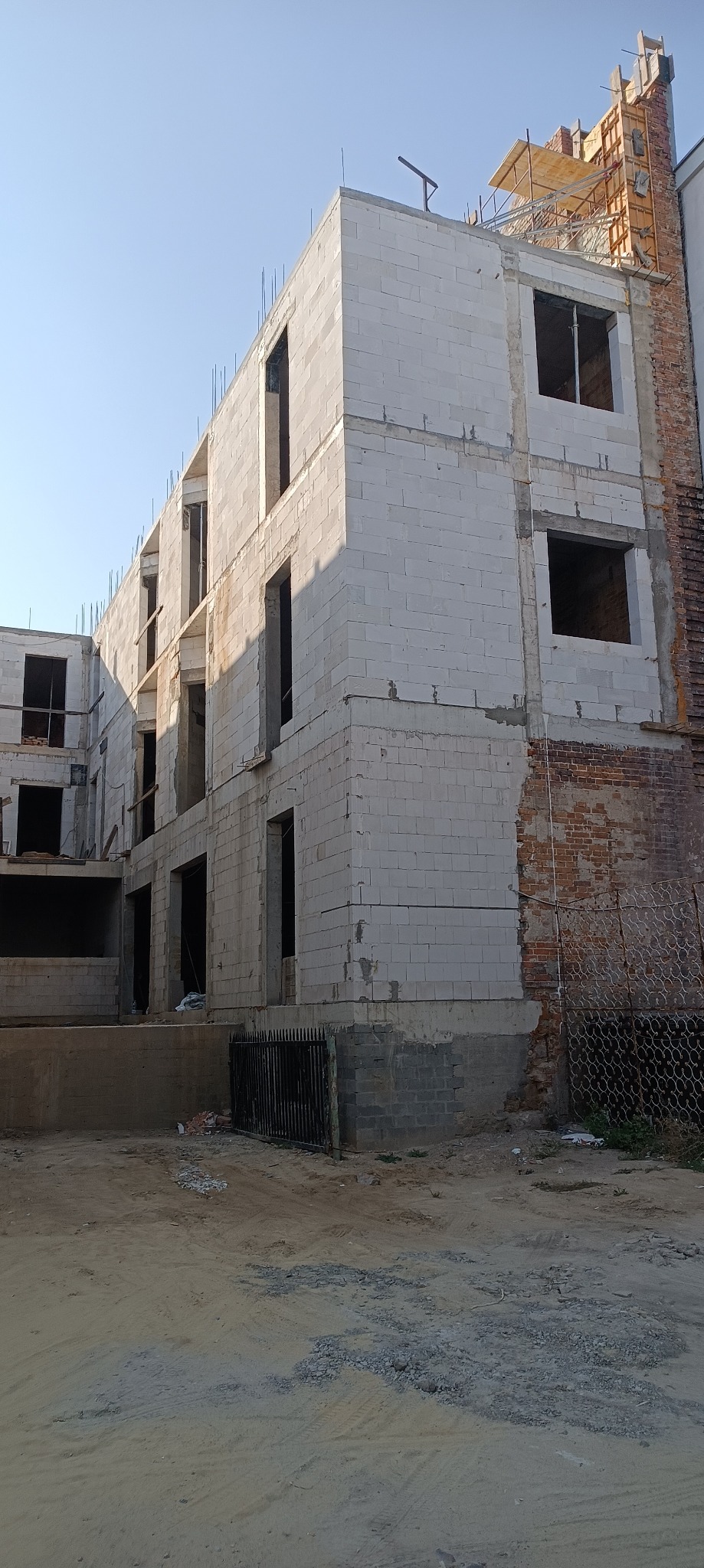 Trzy kondygnacje budynku w stanie surowym otwartym, wzniesione z białych bloczków, z widocznymi otworami okiennymi i elementami szalunków na dachu.