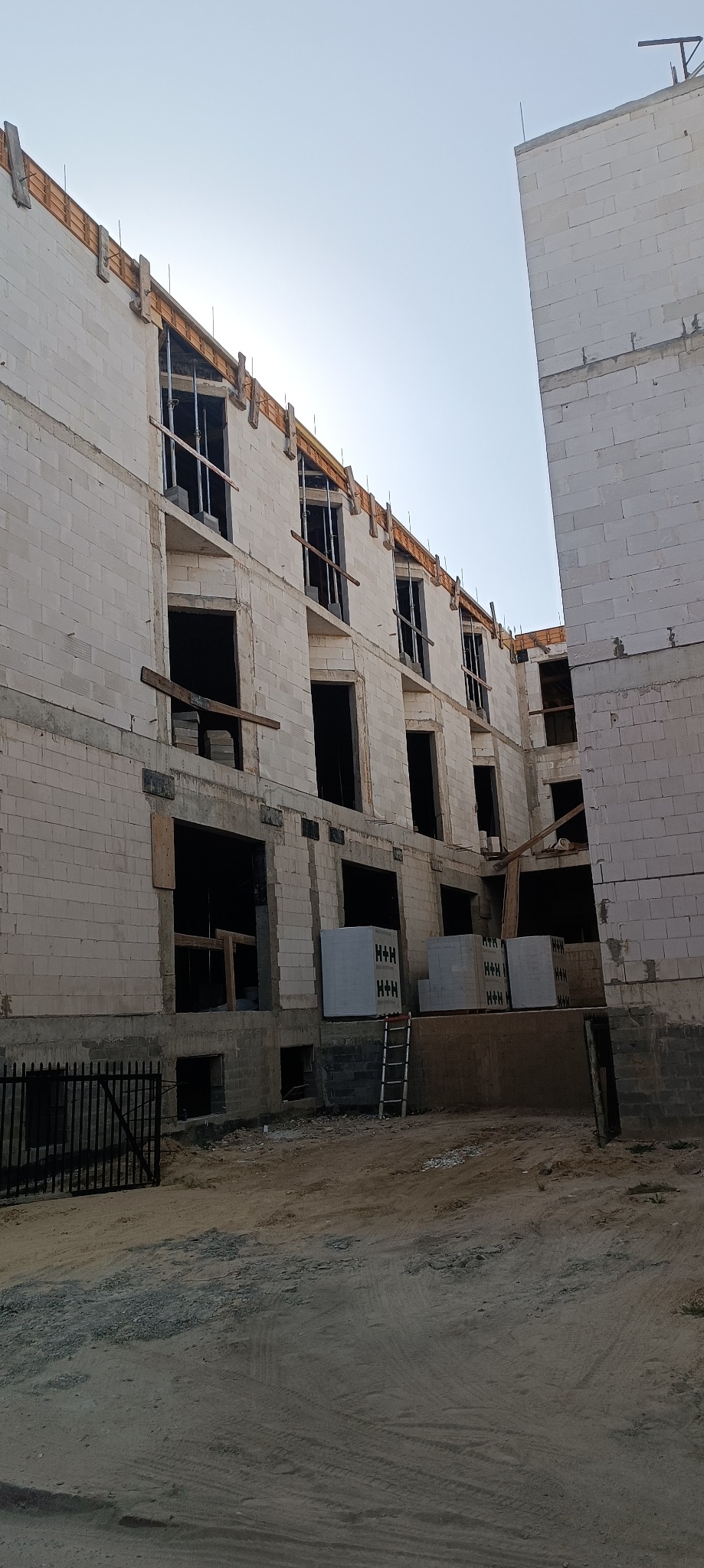 Wznoszenie trzypiętrowego budynku z białych bloczków, widoczne otwarte okna i drewniane szalunki na krawędziach stropów, palety z bloczkami H+H przy ścianie.